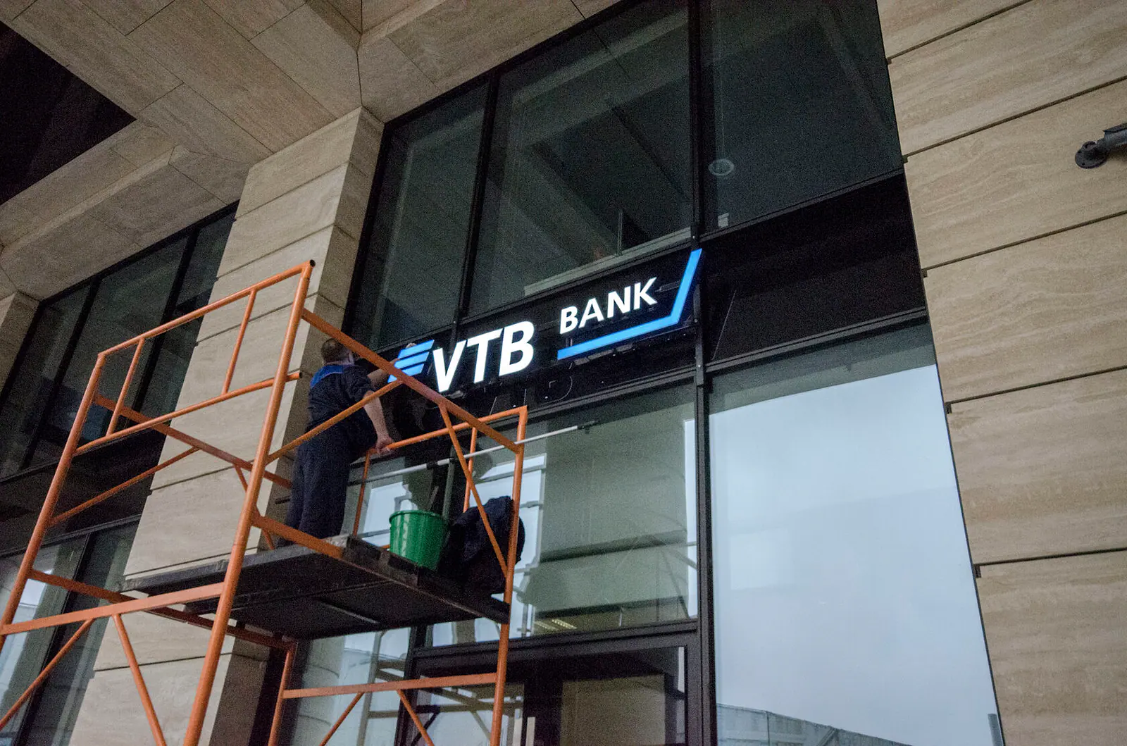 Рабочий на передвижной вышке-туре моет витраж под вывеской VTB Bank